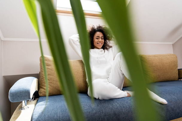 Selektiver Fokus einer glücklichen Afroamerikanerin, die auf einem Sofa in der Nähe einer Pflanze sitzt - Foto, Bild