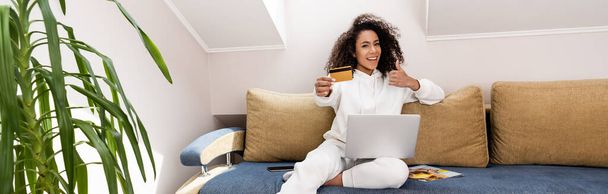 panoramic concept of happy african american girl showing thumb up and holding credit card near laptop  - 写真・画像
