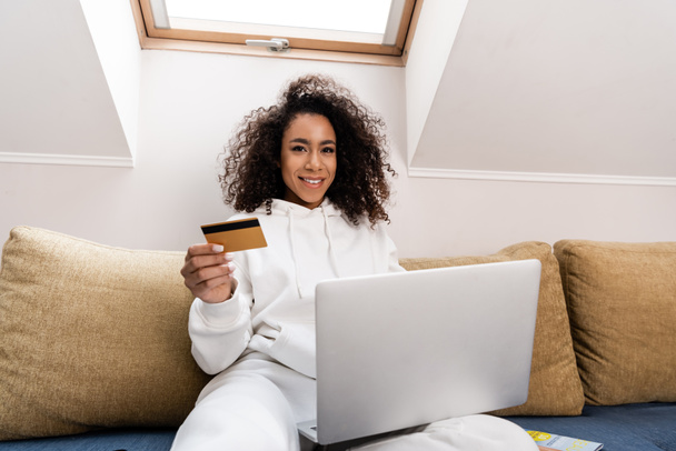 幸せなアフリカ系アメリカ人の女の子ソファに座っている間にラップトップの近くにクレジットカードを保持 - 写真・画像