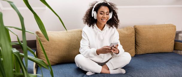 πανοραμικός προσανατολισμός του χαρούμενου Αφροαμερικανού κοριτσιού με ασύρματα ακουστικά χρησιμοποιώντας smartphone ενώ κάθεται στον καναπέ - Φωτογραφία, εικόνα