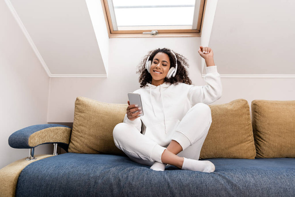 heureuse fille afro-américaine dans un casque sans fil en utilisant un smartphone tout en étant assis sur le canapé
 - Photo, image