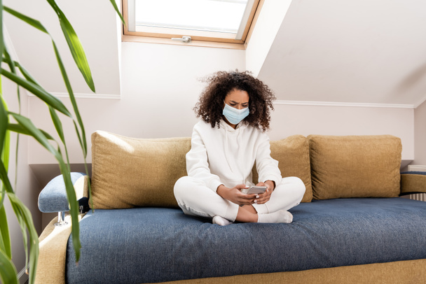 selective focus of curly african american girl in medical mask using smartphone in living room  - Zdjęcie, obraz