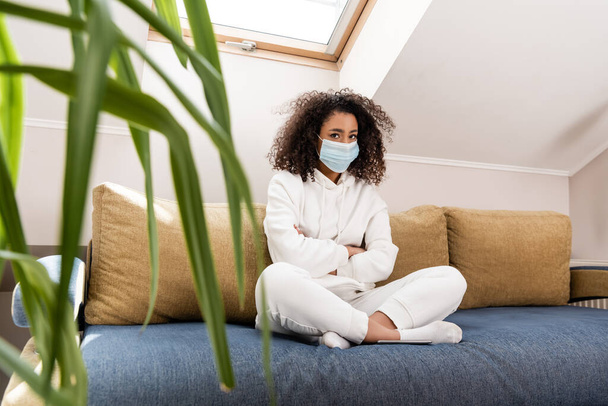 selective focus of curly african american girl in medical mask sitting with crossed arms on sofa  - 写真・画像