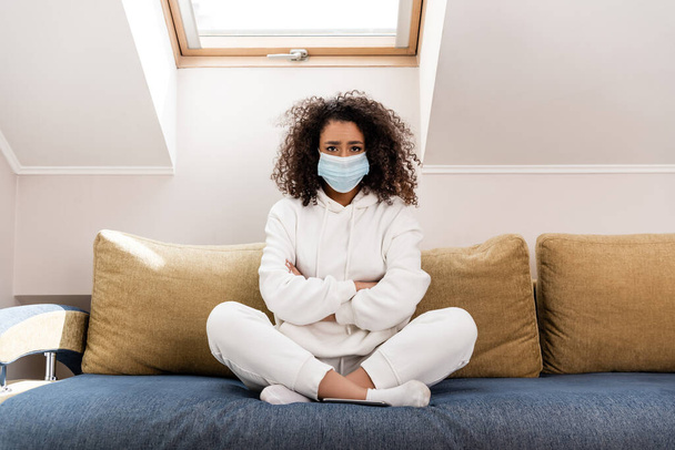 curly african american girl in medical mask sitting with crossed arms on sofa - 写真・画像