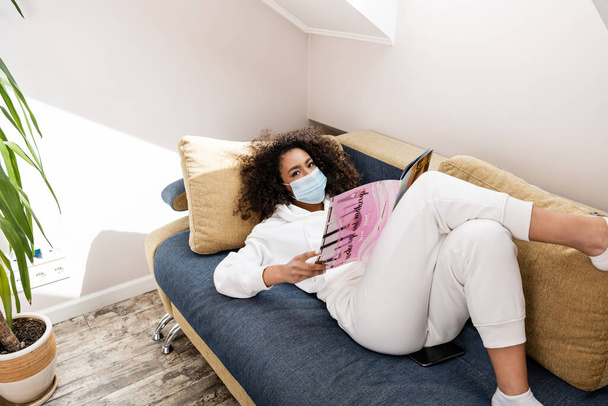 młoda Afroamerykanka w masce medycznej leżąca na kanapie i czytająca magazyn  - Zdjęcie, obraz