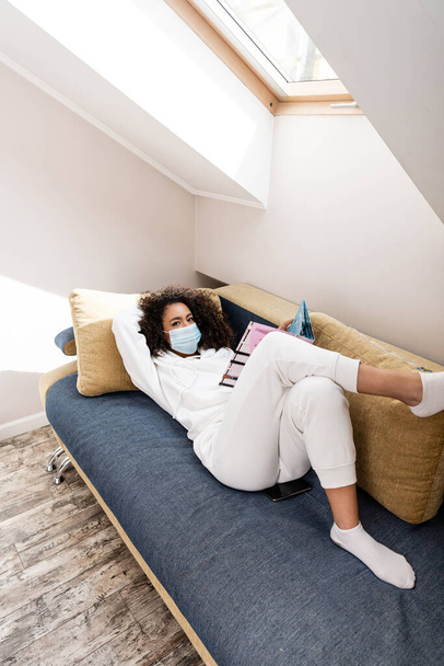 Afrykańska amerykanka w masce medycznej leżąca na kanapie i czytająca magazyn  - Zdjęcie, obraz