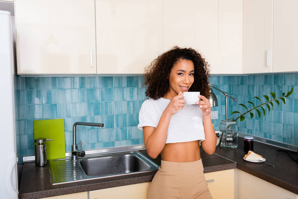happy african american girl looking at camera and holding cup of coffee  - Foto, Imagen