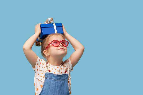 Advertising baby products. Portrait of surprised excited child girl with gift box. isolated over blue background. Empty space for text - Foto, afbeelding