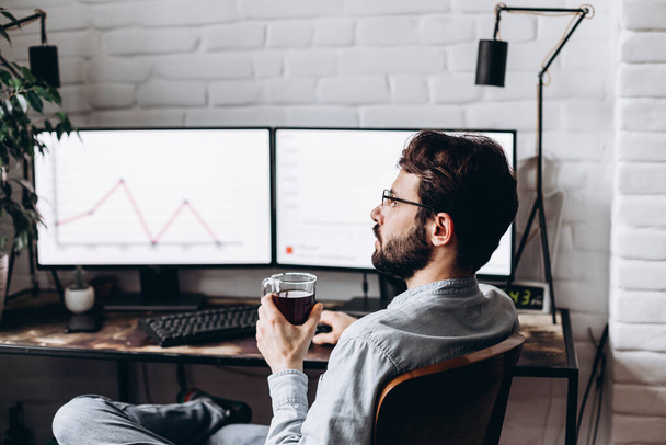 Focused young businessman in eyewear checks graphics on computer monitors. Concentrated millennial man in glasses works at home on self-isolation - Photo, Image