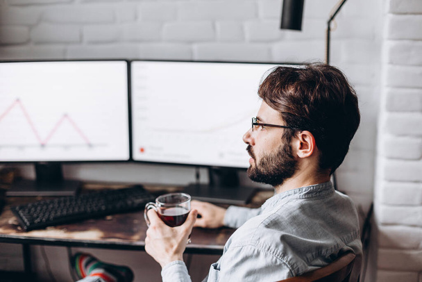 Focused young businessman in eyewear checks graphics on computer monitors. Concentrated millennial man in glasses works at home on self-isolation - Φωτογραφία, εικόνα