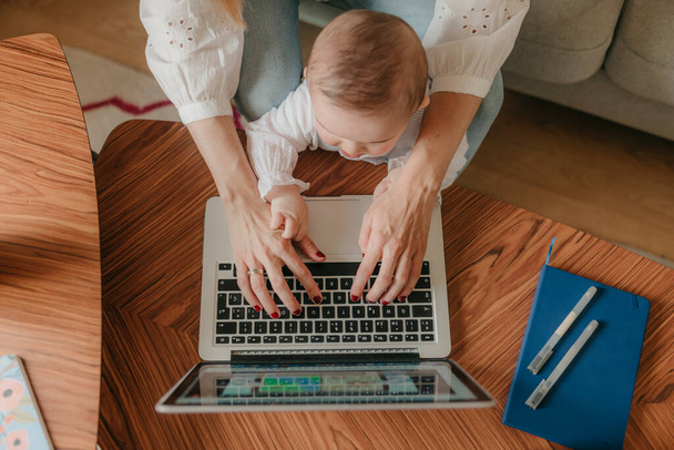 joven madre de negocios está utilizando un ordenador portátil y sonriendo mientras pasa tiempo con su lindo bebé en casa
 - Foto, Imagen