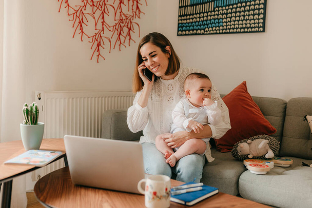 young business mother is using a laptop and smiling while spending time with her cute baby at home - Foto, Imagen
