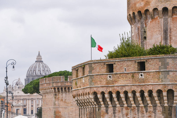 Обзор стен замка Сантанджело и верхней части собора Сан-Пьетро на заднем плане с итальянским флагом
 - Фото, изображение