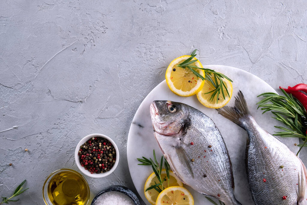Due pesci orata crudi pronti da cuocere con erbe aromatiche, limone e olio d'oliva su tavola di marmo su fondo grigio. Con spazio per la copia. Vista dall'alto
 - Foto, immagini
