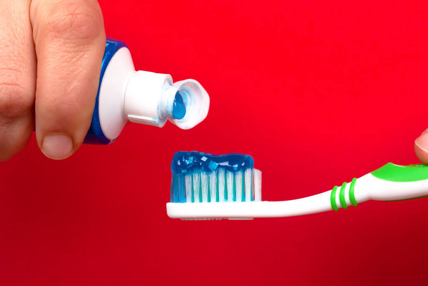 Ruka stiskne zubní pastu na zubním kartáčku na červeném pozadí zblízka. caries prevence a ústní péče - Fotografie, Obrázek