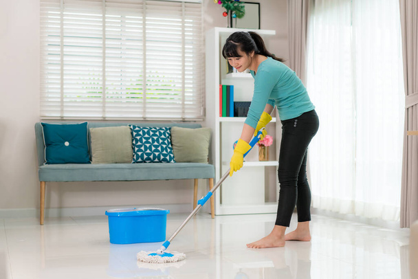 Atractiva joven mujer asiática fregando piso de baldosas en la sala de estar mientras hace la limpieza en casa durante quedarse en casa utilizando el tiempo libre sobre su rutina diaria de limpieza
 - Foto, Imagen
