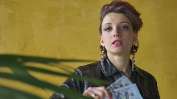Glamouröse aggressive Dame in Punk-Rock-Party-Kleidung mit dunkelrosa Haaren in schwarzer Lederjacke und langen Ohrringen auf gelbem Hintergrund mit Geld-Dollars und Griwna in den Händen - Filmmaterial, Video