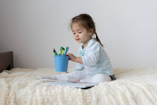 Ein kleines Mädchen europäischen Aussehens sitzt zu Hause auf dem Bett. Sie nahm einen Korb mit Schreibwaren, sie will mit bunten Filzstiften zeichnen. - Foto, Bild