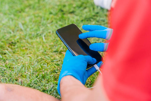 Sportovní chlapec kontroluje svůj mobilní telefon sedí na trávě v parku a poslouchá hudbu v modrých latexových rukavicích pro preventivní opatření potřebná pro koronavirus. - Fotografie, Obrázek