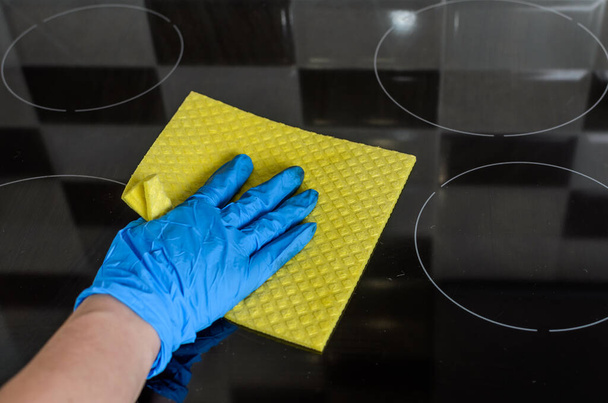 Housekeeper in protective gloves washes the induction cooker with a yellow rag - Foto, afbeelding