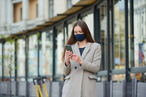 A girl with long hair in a navy blue face mask to avoid the spread coronavirus uses a smartphone in the street. A woman in face mask against COVID-19 wears a coat reads news on a cellphone in the city - Photo, image