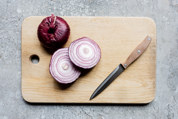 top view of onion on wooden cutting board with knife on grey concrete surface - Fotó, kép