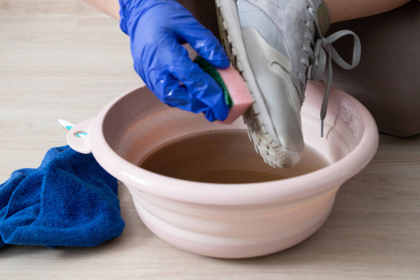 Schmutziges Schuhwerk reinigen und waschen. Frauenhände in blauen Gummi-Latex-Handschuhen halten Waschlappen und Schuhe auf einem Wasserbecken. - Foto, Bild