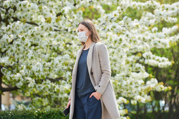 A girl in a medical face mask to avoid the spread coronavirus holds a smartphone in the park. A woman in a face mask against COVID-19 keeps a social distance in the garden between flowering trees. - Foto, imagen
