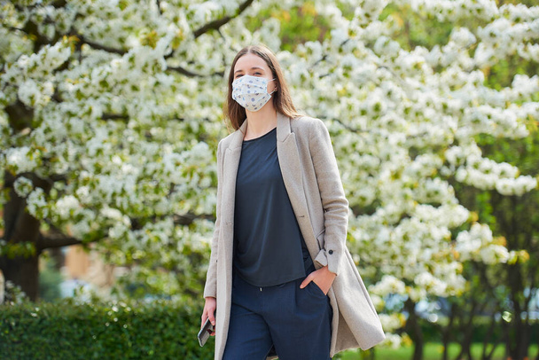 A girl in a medical face mask to avoid the spread coronavirus holds a smartphone in the park. A woman in a face mask against COVID-19 keeps a social distance in the garden between flowering trees. - Foto, imagen