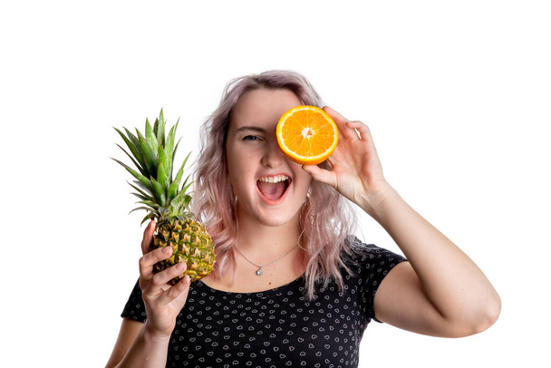 Blond girl with crazy positive emotions in black blouse with a pineapple and orange in her hands. The concept of healthy eating is vegan and vegetarian. Isolated on a white background - Fotó, kép