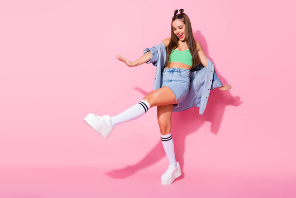 Full body profile photo of funky lady good mood spread hands raise leg hip dancing wear denim jacket green crop top naked belly skirt knee socks shoes isolated pastel pink color background - Photo, image