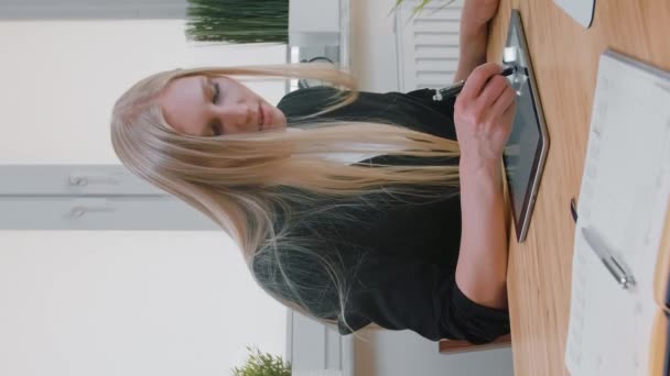 Mulheres de negócios trabalhando em tablet no escritório. Mulher loira atraente com cabelos longos em terno elegante sentado na mesa de madeira com computador e concentrando-se no trabalho em tablet com caneta em escritório leve
. - Filmagem, Vídeo