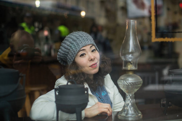Ασιάτισσα που ποζάρει για φωτογράφο με αντανάκλαση στο τζάμι, κοιτάζει πίσω από το παράθυρο της καφετέριας. - Φωτογραφία, εικόνα