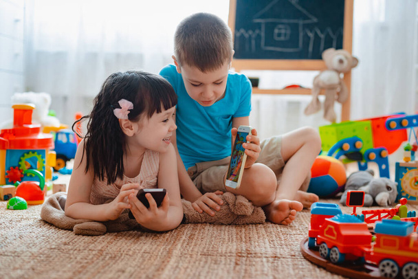 Siblings children brother sister, friends sit floor house children's play room with smartphones, detached scattered toys. Concept new gadgets for kids. Russia. Moscow 30 April 2020 - Фото, зображення