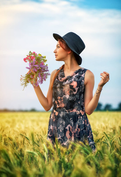 Jolie fille mignonne avec un bouquet de fleurs colorées dans ses mains. La jeune femme respire l'odeur des plantes sur le champ de blé pendant le coucher du soleil. Un regard pensif. Ambiance romantique
. - Photo, image
