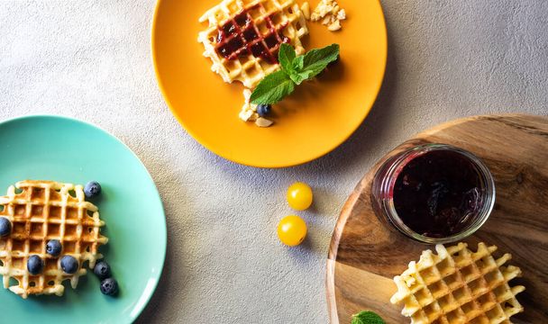 Jälkiruokavohveli täynnä hilloa ja mustikkaa, karpaloa ja keltaisia hedelmiä appelsiinilla ja vihreällä lautasella puupöydällä.
 - Valokuva, kuva