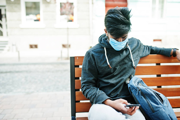 Coronavirus covid-19 concept. South asian indian man wearing mask for protect from corona virus sitting on bench with mobile phone. - Фото, зображення