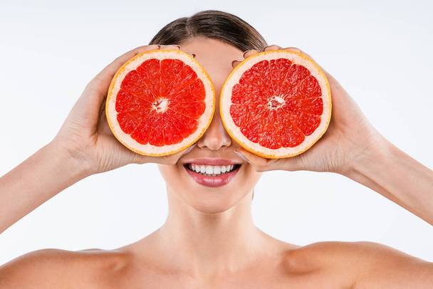 Smiling young woman posing isolated over white wall background holding grapefruit covering eyes - Foto, afbeelding