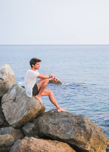 Jeune homme caucasien assis sur un rocher dans une jetée de pierre regardant vers la mer Méditerranée et réfléchissant par temps clair
 - Photo, image