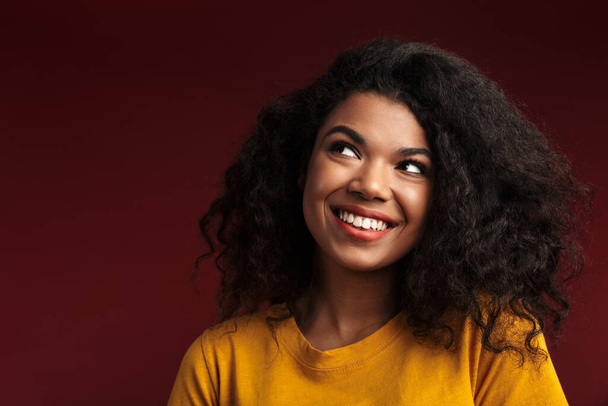 Image of beautiful brunette african american woman with curly hair smiling at camera isolated over red background - 写真・画像