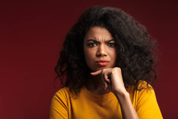 Image of beautiful brunette african american woman with curly hair thinking and touching her chin isolated over red background - Φωτογραφία, εικόνα