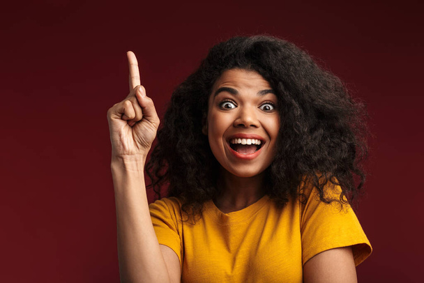 Imagen de la hermosa morena afroamericana con el pelo rizado sonriendo y apuntando con el dedo hacia arriba aislado sobre fondo rojo
 - Foto, imagen
