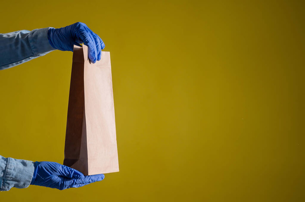 Closeup of female hands in gloves and a denim shirt. Delivery man holds an empty paper bag on a yellow background. Craft packaging for takeaway snack. - 写真・画像