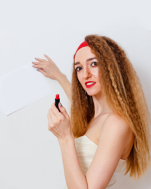 Beautiful young woman with bright makeup and red lipstick in her hands holds a white blank sheet of writing paper on a white background - Fotó, kép