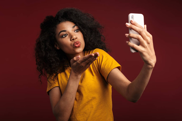 Image of beautiful brunette african american woman sending air kiss while taking selfie photo on cellphone isolated over red background - Fotoğraf, Görsel