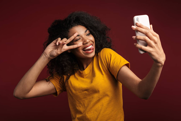 Image of beautiful brunette african american woman gesturing peace fingers while taking selfie photo on cellphone isolated over red background - Φωτογραφία, εικόνα