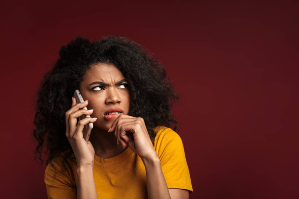 Image of displeased brunette african american woman with curly hair expressing outrage while talking on cellphone isolated over red background - Foto, Imagem