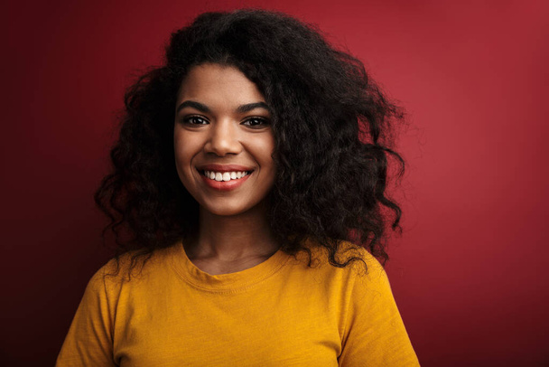 Bild der schönen brünetten afrikanisch-amerikanischen Frau mit lockigem Haar lächelt in die Kamera isoliert über rotem Hintergrund - Foto, Bild