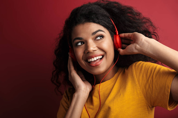 Photo of happy cheery young african woman posing isolated over dark red wall background listening music with headphones. - Foto, Imagem