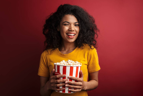 Image of beautiful brunette african american woman with curly hair smiling and holding popcorn bucket isolated over red background - Foto, afbeelding